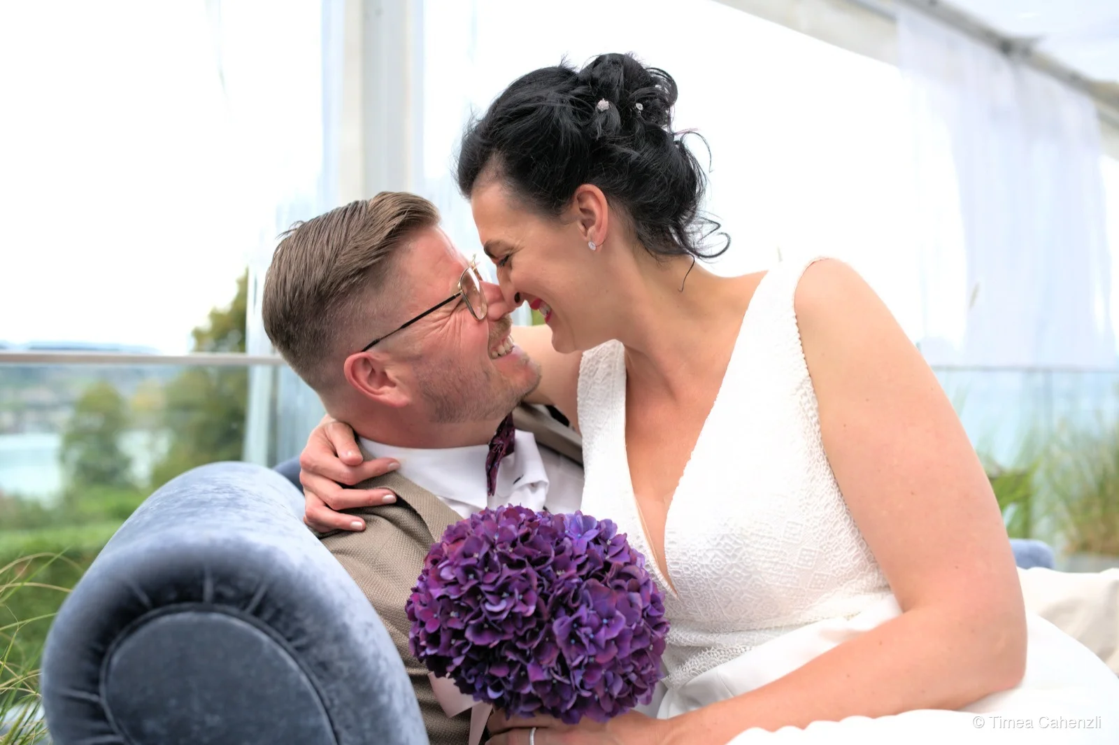 Brautpaar auf Lounge-Sofa – Braut lacht mit lila Hortensienstrauss